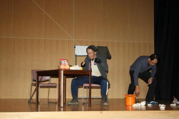 舞台の上でテーブルに座っている男性が散らしたごみを別の男性がごみ箱へ入れようとしている写真